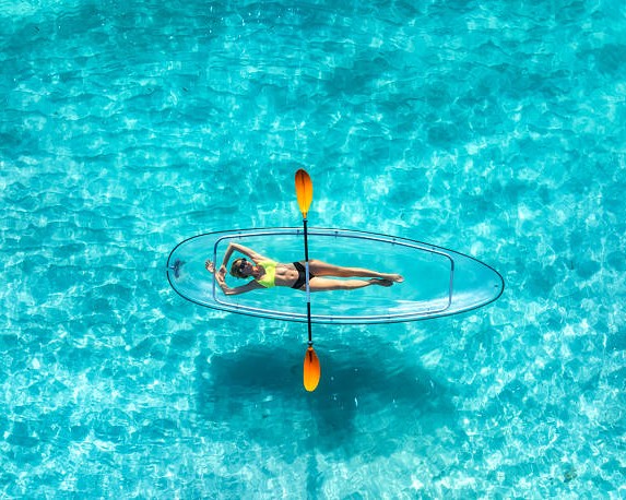 Aerial view of young woman lying on transparent canoe in blue sea at sunrise. Summer in Zanzibar. Top drone view of beautiful slim girl, kanoe, sandy beach, ocean with clear turquoise water. Tropical